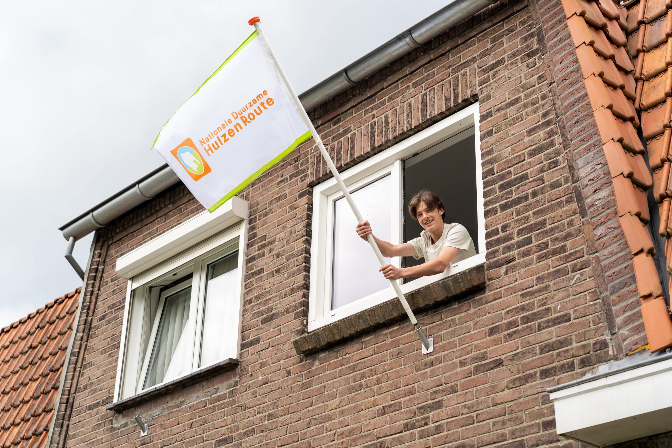 Jongen hangt uit het raam van een woning met de vlag van de Nationale Duurzame Huizen Route